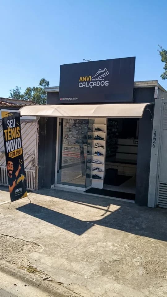 A fachada da Anvi Calçados: a placa preta com a logo, o toldo bege, a vitrine de vidro cheia de tênis e o banner 'Seu tênis novo está aqui!' na calçada