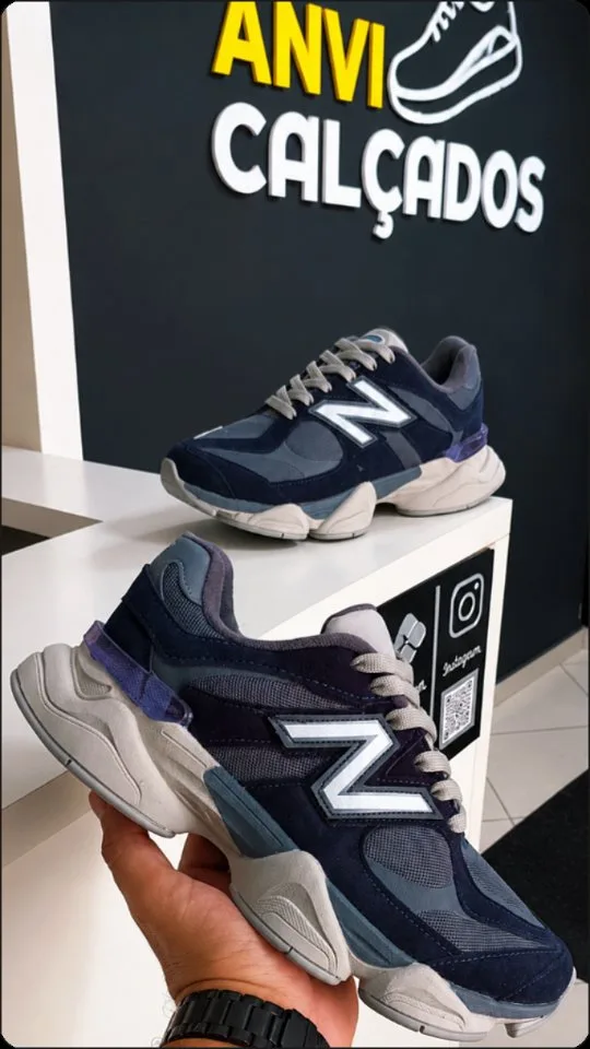 O New Balance 9060 azul-marinho na mão, na frente da parede com a logo da loja